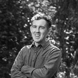 Black and white head and shoulders photograph of Joe Burkinshaw smiling, with a forest behind him