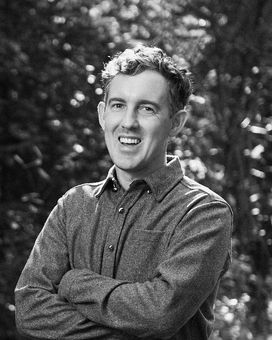 Black and white head and shoulders photograph of Joe Burkinshaw smiling, with a forest behind him